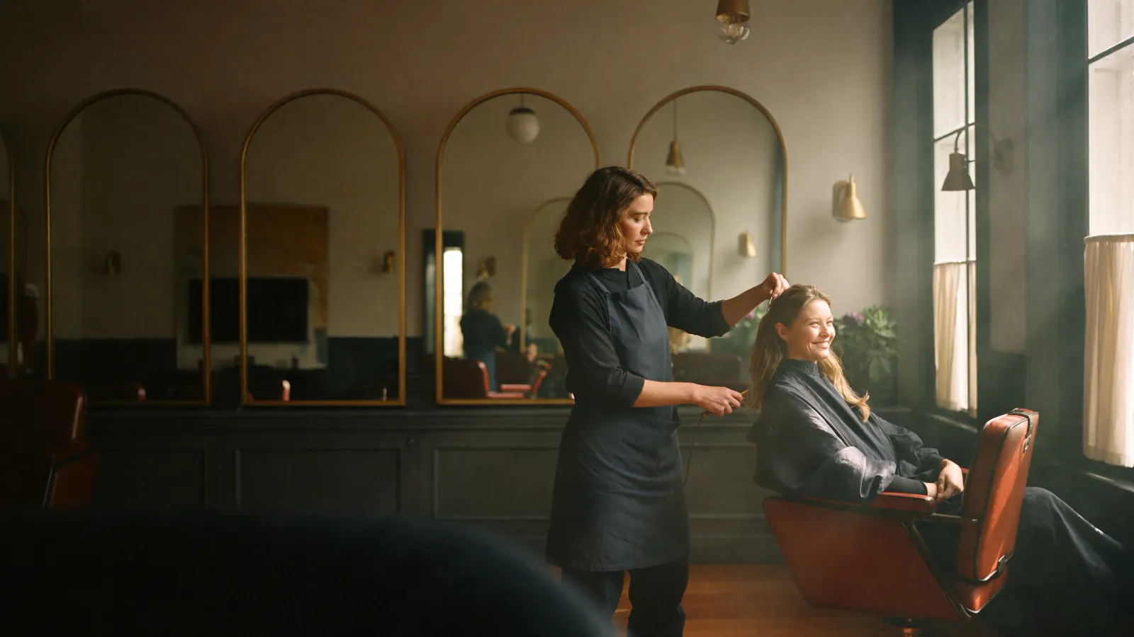 Hairdresser cutting a client's long hair in the sunlit salon with arched mirrors